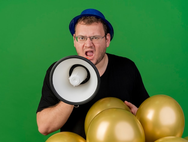 Bezpłatne zdjęcie zirytowany dorosły słowiański mężczyzna w okularach optycznych w niebieskim kapeluszu imprezowym trzyma balony z helem i krzyczy do głośnika