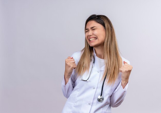 Zirytowany i sfrustrowany młody piękny lekarz kobieta ubrana w biały fartuch z pięściami zaciskającymi stetoskop