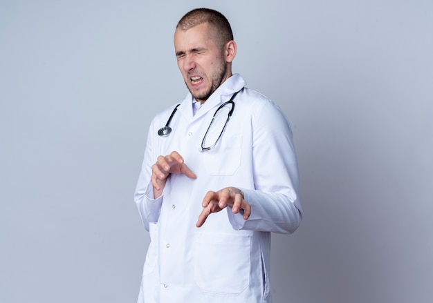 Zirytowany młody lekarz płci męskiej ubrany w szlafrok medyczny i stetoskop na szyi, rozciągający dłoń z zamkniętymi oczami odizolowany na białym z miejscem na kopię