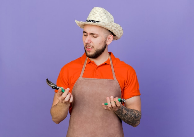 Bezpłatne zdjęcie zirytowany ogrodnik mężczyzna w kapeluszu ogrodniczym trzyma i patrzy na narzędzia ogrodnicze