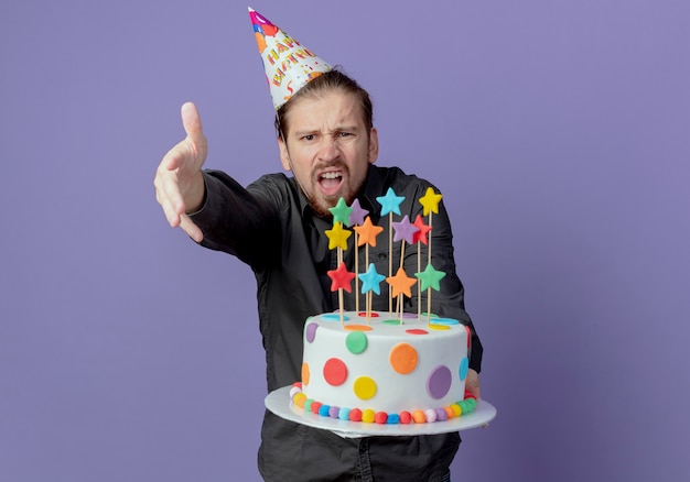Zirytowany przystojny mężczyzna w czapce urodzinowej trzyma tort i wskazuje do przodu ręką odizolowaną na fioletowej ścianie