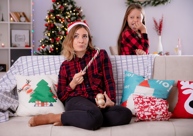 Zła córka zjada część cukierkowej laski, patrząc na matkę w czapce mikołaja trzymającej część złamanej laski i szklanych ozdób w kształcie kulek, siedząc na kanapie w czasie świąt Bożego Narodzenia w domu