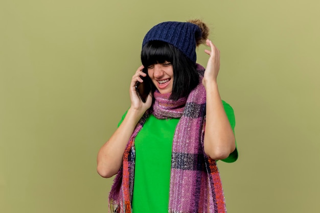 Bezpłatne zdjęcie zła młoda chora kobieta w czapce zimowej i szaliku rozmawia przez telefon trzymając rękę w powietrzu z zamkniętymi oczami odizolowaną na oliwkowej ścianie z miejscem na kopię