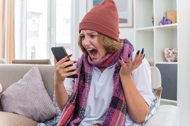 Zła niezdrowa młoda kobieta w kapeluszu z ciepłym szalikiem na szyi źle się czuje i choruje cierpi na przeziębienie i grypę krzyczy podczas rozmowy przez telefon komórkowy, siedząc na kanapie w jasnym salonie