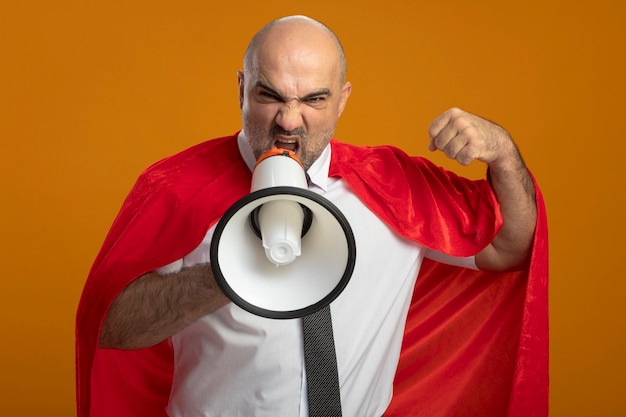 Bezpłatne zdjęcie zły biznesmen super bohater w czerwonej pelerynie krzyczy do megafonu, zaciskając pięść stojącą nad pomarańczową ścianą