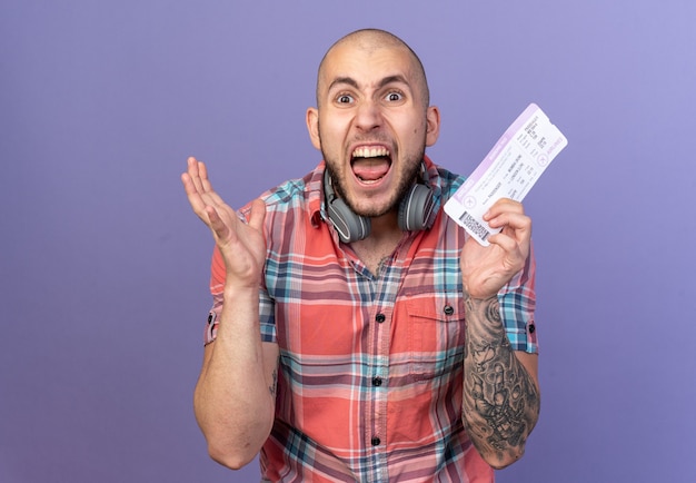 Bezpłatne zdjęcie zły młody podróżnik ze słuchawkami na szyi trzymający bilet lotniczy odizolowany na fioletowej ścianie z kopią miejsca
