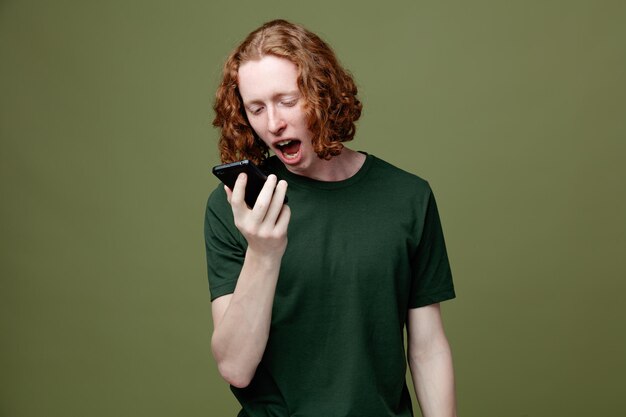 Zły młody przystojny facet trzymający i patrzący na telefon ubrany w zieloną koszulkę na białym tle na zielonym tle