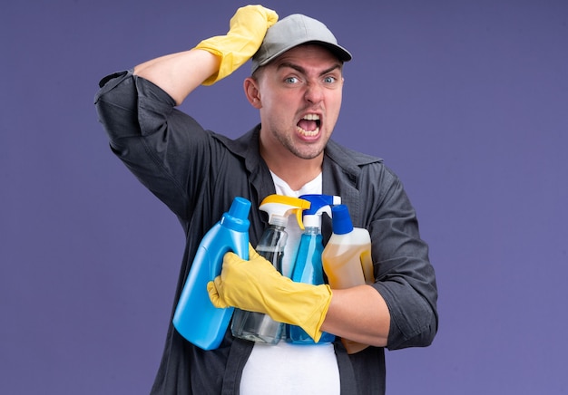 Zły młody przystojny sprzątacz sobie t-shirt z czapką i rękawiczkami, trzymając narzędzia do czyszczenia, kładąc rękę na głowie na białym tle na fioletowej ścianie