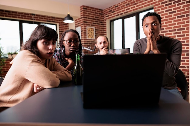 Bezpłatne zdjęcie zmartwieni wieloetniczni przyjaciele siedzą w domu w salonie, oglądając tragiczne wydarzenia na nowoczesnym laptopie. różnorodni ludzie siedzą przy stole podczas wspólnego oglądania horrorów na komputerze.