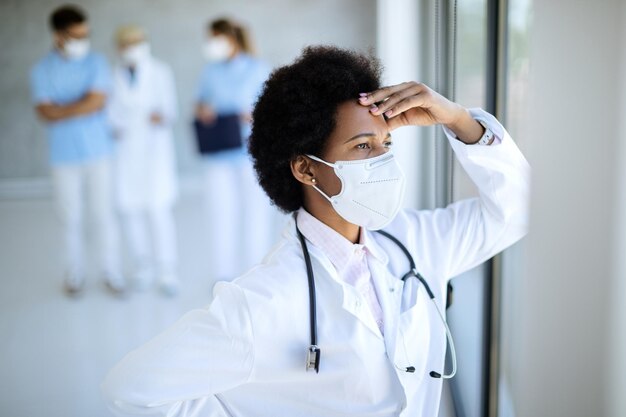 Zmartwiona Afroamerykanka lekarka z maską na twarz patrząca przez okno