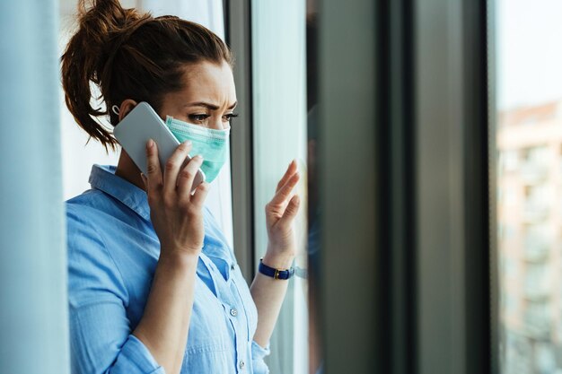 Zmartwiona kobieta nosi maskę na twarz, komunikując się przez telefon komórkowy i patrząc przez okno