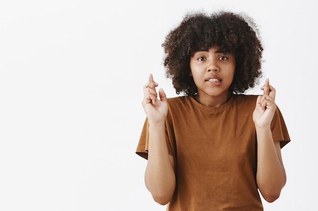 Zmartwiona niepewna, atrakcyjna kobieta o ciemnej karnacji z uroczą fryzurą w stylu afro zaciskająca zęby, trzymająca palce na szczęście, modląc się o zdanie egzaminu z dobrym wynikiem, aby otrzymać stypendium