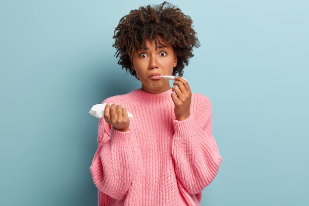 Zmartwiona piękna niezdrowa kobieta ma przeziębienie lub gorączkę, mierzy temperaturę, trzyma chusteczkę, zaskoczona grypą, nosi za duży różowy sweter, trzyma termometr w ustach, odizolowany na niebieskiej ścianie.