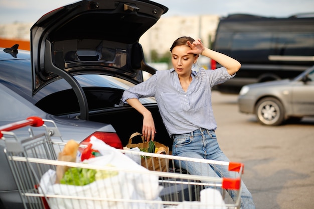 Zmęczona kobieta z wózkiem wkłada swoje zakupy do bagażnika samochodu na parkingu supermarketu. zadowolony klient niosący zakupy z centrum handlowego, pojazdy w tle