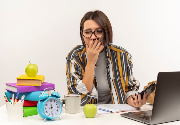 Zmęczona młoda studentka w okularach siedzi przy biurku z narzędziami uniwersyteckimi ziewająca z ręką na ustach i zamkniętymi oczami i trzymając telefon komórkowy na białym tle