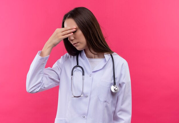 Zmęczony młody lekarz dziewczyna ubrana w stetoskop medycznej sukni kładąc rękę na nosie na na białym tle różowej ścianie