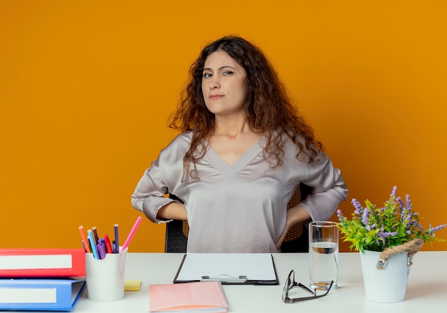 Bezpłatne zdjęcie zmęczony pracownik biurowy młodych całkiem żeński siedzi przy biurku z narzędzi biurowych, kładąc ręce na plecach na białym tle na pomarańczowy