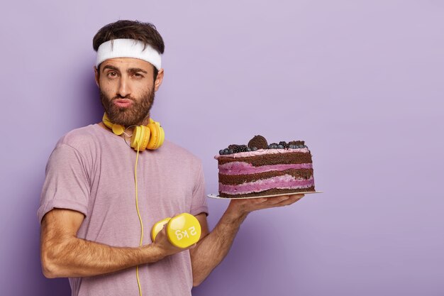 Bezpłatne zdjęcie zmotywowany poważny facet ma mocne bicepsy, trzyma żółte hantle, prowadzi zdrowy tryb życia, trzyma świeżo upieczone ciasto