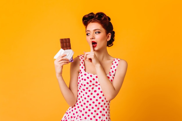 Zmysłowa kobieta pinup jedzenie czekolady na żółtej przestrzeni. Imbirowa dziewczyna w sukience w kropki dotykając ust palcem.