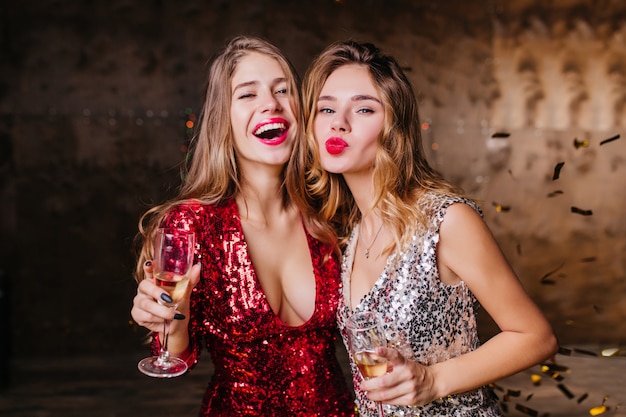 Zmysłowa kobieta w czerwonej modnej sukience szczęśliwa śmiejąc się, gdy jej koleżanka pozuje z całowaniem wyrazem twarzy