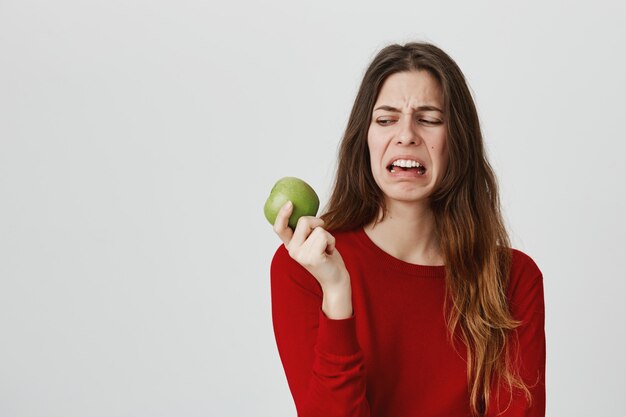 Zniesmaczona śliczna kobieta patrzy na jabłko z niechęcią i niechęcią, krzywiąc się