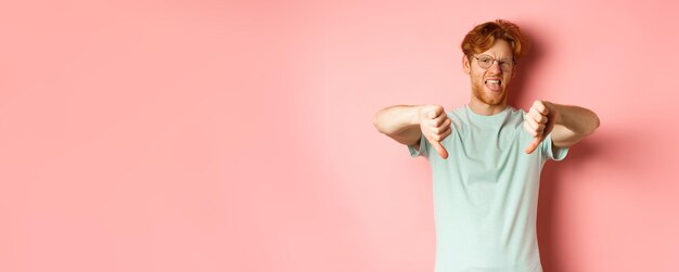 Bezpłatne zdjęcie zniesmaczony rudy mężczyzna w koszulce i okularach, pokazujący kciuki w dół i język krzywiący się z niechęci