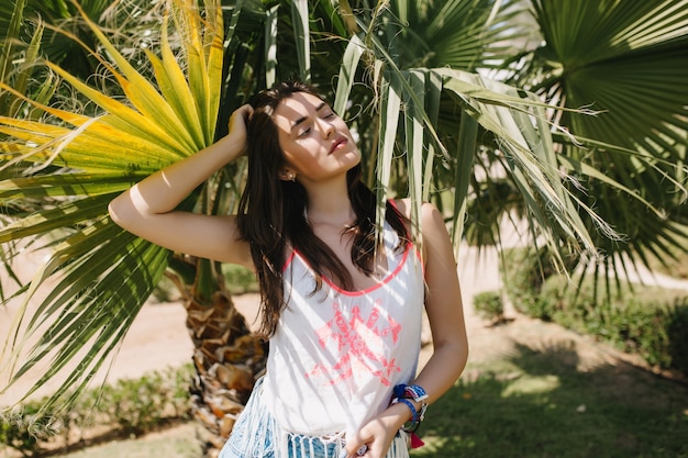 Zniewalająca dziewczyna o ciemnych prostych włosach odpoczywa w cieniu palm na wakacjach w egzotycznym kraju. Portret ładny młoda kobieta pozuje z zamkniętymi oczami z południowych roślin