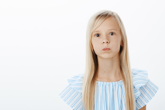 Znudzona i beztroska mała dziewczynka próbuje rozweselić, wygłupia się. Portret figlarnego uroczego dziecka płci żeńskiej o blond włosach, dąsa się, wstrzymuje oddech i wpatruje się w wytrzeszczone oczy