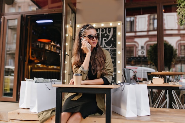 Znudzona kobieta dzwoni do kogoś siedząc w kawiarni na świeżym powietrzu po zakupach