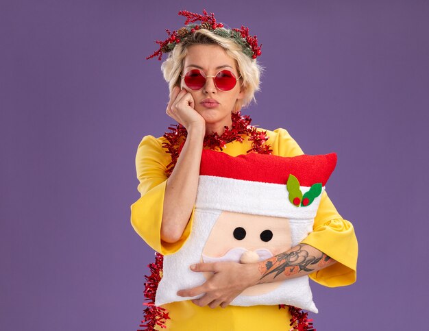 Znudzona młoda blondynka ubrana w świąteczny wieniec na głowę i świecącą girlandę wokół szyi, trzymając poduszkę świętego mikołaja, trzymając rękę na twarzy, patrząc na kamerę odizolowaną na fioletowym tle