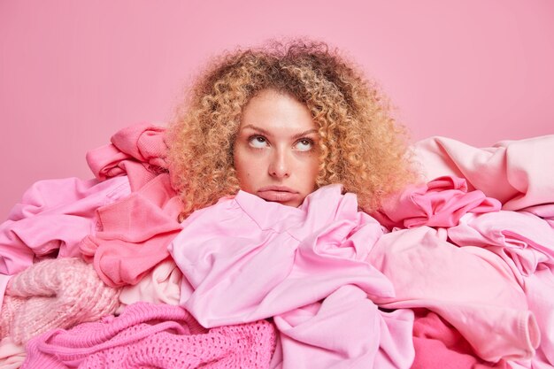 Znudzona, smutna, młoda kobieta z kręconymi, krzaczastymi włosami pozuje wokół stosów ubrań skupionych ponad na tle różowej ściany. Zaśmiecona kobieca garderoba. Recykling tkanin i koncepcja ponownego wykorzystania tekstyliów.