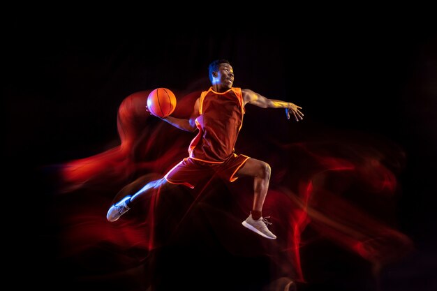 Zobacz cel. Afro-koszykarz młody zespół czerwony w akcji i neony na ciemnym tle studio. Pojęcie sportu, ruchu, energii i dynamicznego, zdrowego stylu życia.