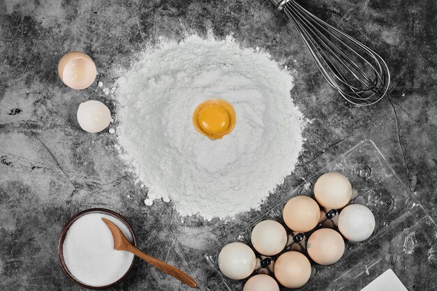 Żółtko na mące, jajka, miska mąki i wąsy na kamiennym stole.