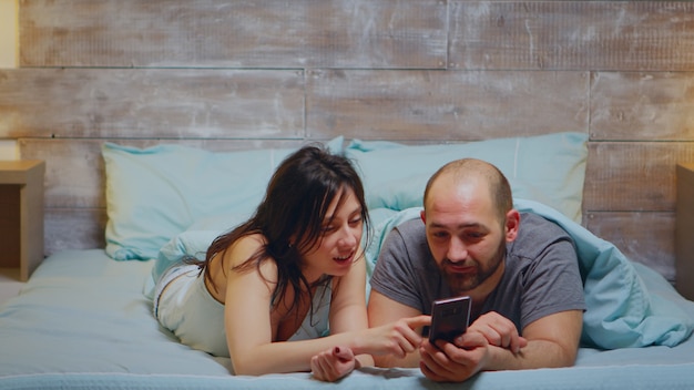 Żona W Piżamie śmiejąca Się, Podczas Gdy Jej Mąż Pokazuje Coś Przez Telefon W Sypialni