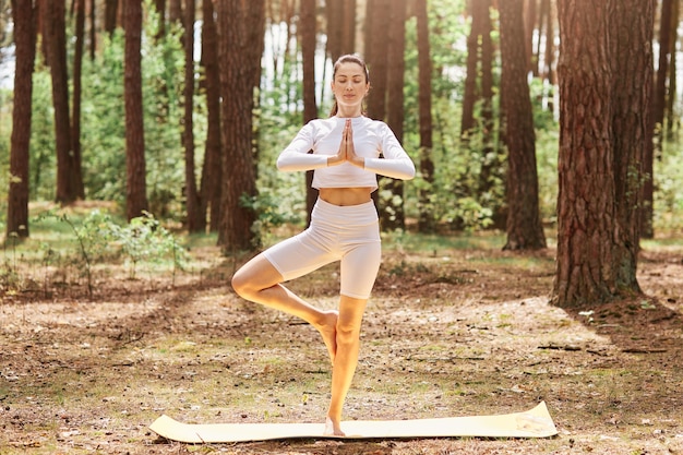 Zrelaksowana wysportowana kobieta stojąca na świeżym powietrzu z zamkniętymi oczami trzymająca dłonie razem, stojąca na jednej nodze, ubrana w sportową odzież, ciesząca się treningiem w pięknym lesie.