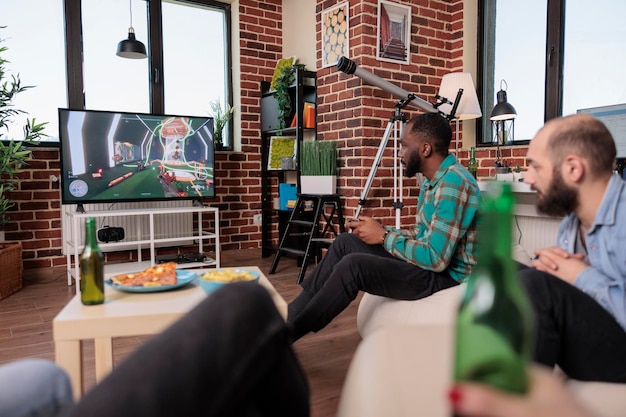 Zróżnicowana grupa ludzi grających w gry wideo na konsoli telewizyjnej, pijących piwo na spotkaniu towarzyskim z przyjaciółmi. Zabawa z rywalizacją w grach i spędzaniem wolnego czasu na imprezie domowej.