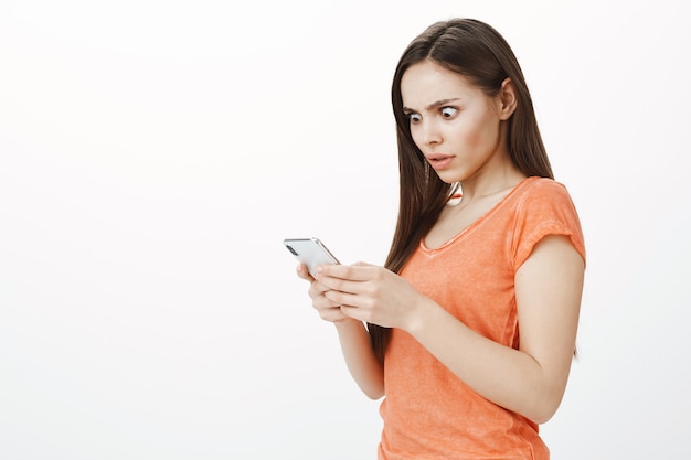Zszokowana atrakcyjna dziewczyna wyskakująca oczy na ekranie, wpatrująca się w wyświetlacz telefonu komórkowego z niespokojnym spojrzeniem
