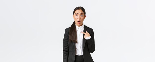 Zszokowana i obrażona młoda azjatycka kobieta w garniturze wskazująca na siebie ze zdumioną paniką na twarzy jest oskarżana Businesswoman patrząc urażoną po tym, jak została nazwana białym tle
