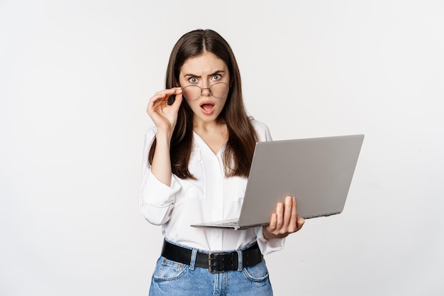 Zszokowana kobieta patrząca na ekran laptopa zdezorientowana, oszołomiona czymś na komputerze, stojąca na białym tle