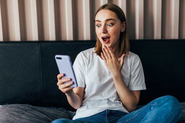 Zszokowana młoda kobieta patrząca na telefon komórkowy w łóżku w domu