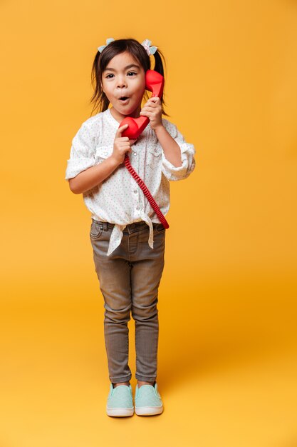 Zszokowana podekscytowana dziewczynka rozmawia przez czerwony telefon retro.