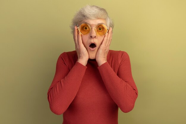 zszokowana stara kobieta w czerwonym swetrze z golfem i okularach przeciwsłonecznych, patrząc na kamerę, kładąc ręce na twarzy odizolowanej na oliwkowozielonym tle z kopią przestrzeni