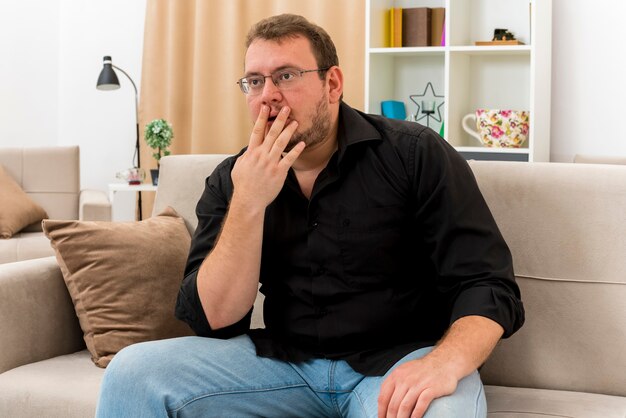 Zszokowany dorosły słowiański mężczyzna w okularach optycznych siedzi na fotelu, kładąc dłoń na ustach i patrząc z boku na wnętrze salonu