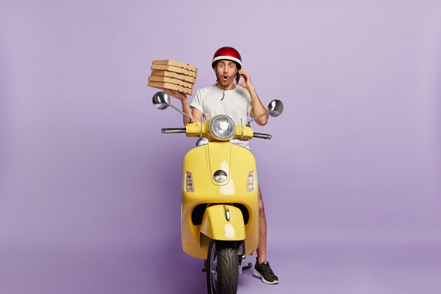 Zszokowany dostawca prowadzący skuter, trzymając pudełka po pizzy