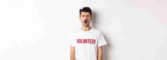 Bezpłatne zdjęcie zszokowany i zdezorientowany mężczyzna w koszulce wolontariusza, wpatrujący się w kamerę i marszczący brwi, niezadowolony, stojący agai