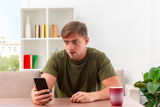 Zszokowany młody przystojny mężczyzna blondynka siedzi przy stole z kubkiem, trzymając i patrząc na telefon w salonie