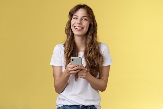 Żywa entuzjastyczna przyjazna uśmiechnięta szczęśliwa kobieta za pomocą smartfona wysyłanie wiadomości tekstowych przyjacielowi sprawdzanie kanałów mediów społecznościowych przeglądanie Internetu trzymaj telefon komórkowy śmiejąc się szczęśliwie żółtym tle.