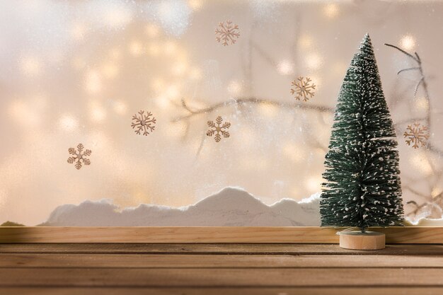 Abeto de juguete en una mesa de madera cerca del banco de nieve, ramita de plantas, copos de nieve y luces de colores