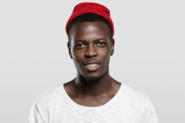 Abierta y sociable. Hermoso hipster afroamericano joven feliz con sombrero rojo de moda, sonriendo, mostrando sus dientes blancos.