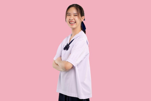 Abrazaderas feliz chica estudiante asiática en uniforme escolar con el brazo cruzado sobre fondo pastel aislado